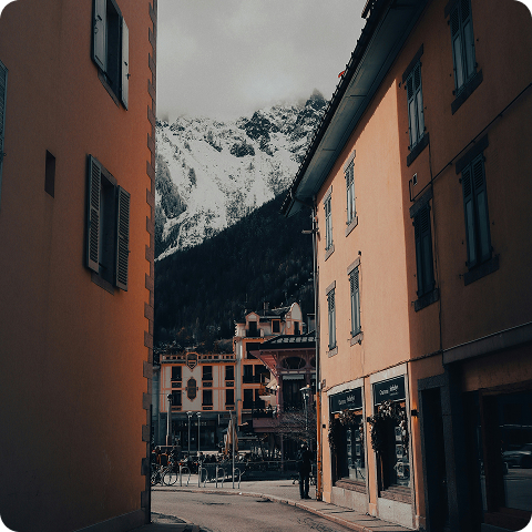 Cette photographie atmosphérique capture l'essence d'une station ou d'un village de montagne où le tissu urbain historique dialogue harmonieusement avec l'environnement naturel spectaculaire. La composition encadrée crée un effet de découverte saisissant. Premier plan - architecture urbaine : Deux bâtiments traditionnels encadrent la perspective : façades aux enduits ocre et saumon caractéristiques de l'architecture alpine italienne ou savoyarde, menuiseries bois peintes (volets et fenêtres) aux teintes vertes ou grises patinées, rez-de-chaussée commerciaux avec vitrines et enseignes (magasin visible à droite "Demi..."), fenêtres à petits carreaux traditionnels typiques de l'architecture montagnarde ancienne, architecture de 3 à 4 niveaux créant une échelle urbaine intime, et détails architecturaux (encadrements, corniches) témoignant du patrimoine bâti préservé. Second plan - place et vie urbaine : La ruelle s'ouvre sur une placette animée : bâtiment central aux façades colorées (rose, ocre) avec rez-de-chaussée commercial, terrasses de cafés ou restaurants visibles avec parasols et mobilier urbain, quelques passants et cyclistes animant l'espace public, fontaine ou monument peut-être présent au centre (difficilement identifiable), et atmosphère conviviale de centre-ville piétonnier ou semi-piétonnier. Arrière-plan - le sommet : Un pic enneigé majestueux ferme spectaculairement la perspective : sommet rocheux acéré culminant probablement entre 2500 et 3500 mètres, versants nord conservant la neige et les glaces même en intersaison, couloirs d'avalanche et combes enneigées striés sur les pentes raides, forêts de conifères sombres couvrant l'étage subalpin, et ciel gris légèrement voilé créant une ambiance crépusculaire ou de fin de journée. Lumière et atmosphère : L'éclairage crée une ambiance particulière : lumière douce et diffuse de fin de journée ou de temps couvert, façades chaudes (ocre, saumon) contrastant avec la froideur minérale du sommet, ombres portées dans la ruelle renforçant la profondeur, atmosphère intimiste et authentique typique des villages alpins préservés, et sentiment de proximité immédiate entre ville et montagne. Composition et cadrage : Le cadrage intelligent renforce l'impact visuel : effet de cadre naturel créé par les bâtiments latéraux, ligne de fuite dirigeant le regard vers le sommet spectaculaire, contraste saisissant entre l'urbain au premier plan et le sauvage à l'arrière-plan, et sentiment de découverte progressive révélant la montagne au bout de la rue.