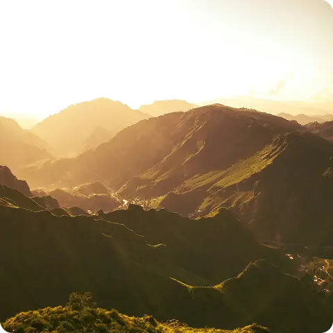Voici une description de cette image : Description courte : Lever ou coucher de soleil doré sur une vallée alpine, révélant l'étagement majestueux des massifs montagneux dans une atmosphère lumineuse exceptionnelle. Description détaillée : Cette photographie atmosphérique capture la magie d'un moment privilégié en montagne, probablement à l'aube ou au crépuscule, lorsque la lumière rasante transforme le paysage alpin en un tableau de couleurs chaudes et de profondeurs infinies. Premier plan - versants et vallée : Les pentes du premier plan plongent dans l'ombre tout en révélant leur structure : versants herbeux aux teintes vert sombre ponctuées par les derniers rayons dorés, prairies d'altitude ondulant selon les courbes naturelles du relief, quelques arbres ou bosquets isolés créant des taches sombres, et chalets ou habitations dispersées minuscules témoignant de la présence humaine discrète. Second plan - les massifs intermédiaires : Une succession de crêtes et de vallées crée une profondeur spectaculaire : montagnes aux silhouettes arrondies caractéristiques de certains massifs alpins ou préalpins, versants striés par les couloirs d'érosion et les ravines, végétation dense (forêts de conifères probablement) couvrant les pentes jusqu'à l'étage alpin, vallée principale se dessinant en contrebas avec brumes légères flottant au fond, et jeu d'ombres et de lumières révélant chaque relief avec une précision sculpturale. Arrière-plan - sommets lointains : L'horizon se perd dans une succession de plans montagneux : chaînes de montagnes s'estompant progressivement dans la brume atmosphérique dorée, sommets culminants aux formes douces se fondant dans le ciel lumineux, dégradé subtil du brun doré au jaune pâle créant une perspective aérienne remarquable, et sentiment d'infini et d'immensité caractéristique des grands massifs alpins. Lumière et atmosphère : L'éclairage crée toute la magie de l'image : lumière rasante dorée du lever ou coucher de soleil illuminant les sommets et les crêtes, ombres longues plongeant les vallées dans une semi-pénombre mystérieuse, atmosphère dorée et vaporeuse créée par la brume légère et les particules en suspension, tonalités chaudes allant du vert doré au brun ambré en passant par l'ocre, et qualité de lumière exceptionnelle caractéristique de la golden hour en montagne. Sentiment et émotion : L'ensemble dégage une sensation de : sérénité et contemplation face à la grandeur de la nature, beauté éphémère et précieuse d'un instant unique, immensité et infini des espaces montagnards, harmonie et équilibre entre lumière, relief et végétation, et privilège d'assister à ce spectacle naturel quotidien mais toujours renouvelé.