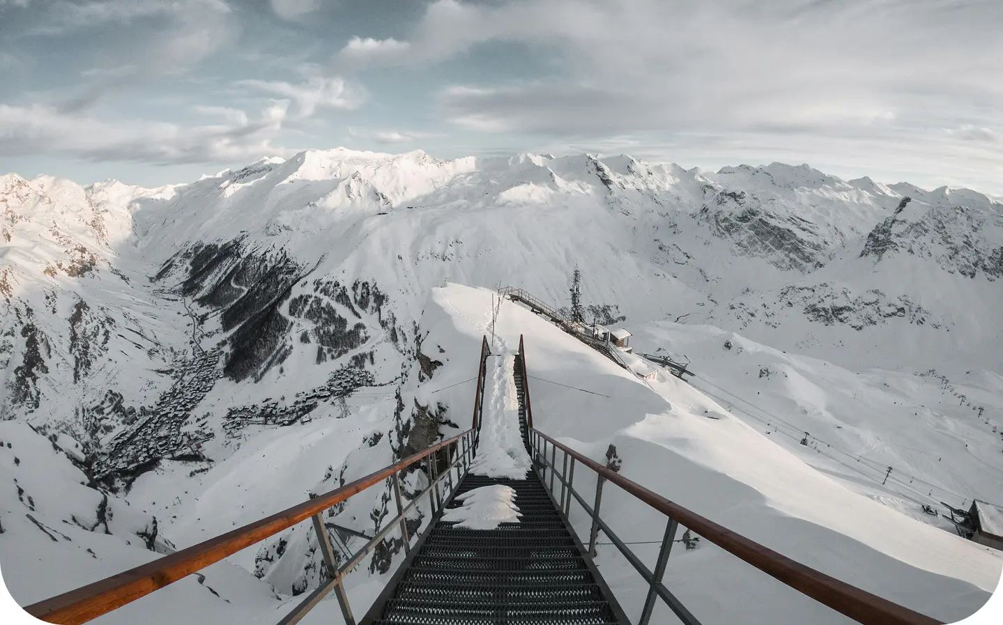 Cette photographie spectaculaire capture la montée vers un restaurant, observatoire ou point de vue d'altitude, illustrant parfaitement l'audace architecturale des équipements de haute montagne et la beauté vertigineuse des Alpes. Premier plan - l'escalier et les infrastructures : Un escalier métallique impressionnant s'élance vers les hauteurs : structure en acier galvanisé ou peint avec marches grillagées antidérapantes, garde-corps tubulaires rouge-orangé apportant une touche de couleur dans l'immensité blanche, construction audacieuse ancrée dans la roche et la neige sur une arête sommitale étroite, perspective plongeante vertigineuse révélant le vide de part et d'autre, et main courante bois offrant une prise chaleureuse et naturelle. Au sommet de l'escalier, on aperçoit un bâtiment d'altitude : construction compacte adaptée aux conditions extrêmes, toiture recouverte de neige fraîche, et antenne ou pylône télécom émergeant du toit. Second plan - l'arête sommitale : Une crête enneigée spectaculaire structure l'espace : arête étroite serpentant entre les versants vertigineux de part et d'autre, corniches neigeuses sculptées par le vent témoignant des conditions météorologiques rigoureuses, installations de remontées mécaniques (pylônes, câbles) visible le long de l'arête, pistes de ski damées dévalant les versants adjacents, et quelques skieurs minuscules donnant l'échelle monumentale du site. Arrière-plan - l'amphithéâtre de sommets : Un panorama montagnard exceptionnel se déploie à 360° : succession de sommets enneigés aux formes variées créant une profondeur infinie, massifs aux altitudes probablement comprises entre 2800 et 3500 mètres, versants alternant pentes douces et faces raides, zones rocheuses affleurant sous la neige révélant la structure géologique, glaciers et névés accrochés aux versants nord, et chaînes montagneuses s'estompant progressivement à l'horizon dans la brume atmosphérique. Ciel et lumière : Les conditions météorologiques créent une atmosphère dramatique : ciel tourmenté avec accumulation de nuages gris menaçants, éclaircies laissant filtrer une lumière diffuse, contrastes subtils de gris, blancs et bleus pâles, atmosphère changeante typique de la haute montagne, et sensation d'immensité et de puissance des éléments naturels. Sensation et émotion : L'image inspire : vertige et fascination face au vide et à l'altitude, admiration pour l'audace technique et architecturale, sentiment de privilège d'accéder à de tels panoramas, respect de la montagne et de ses dangers, et émerveillement devant la beauté sauvage et préservée des Alpes.