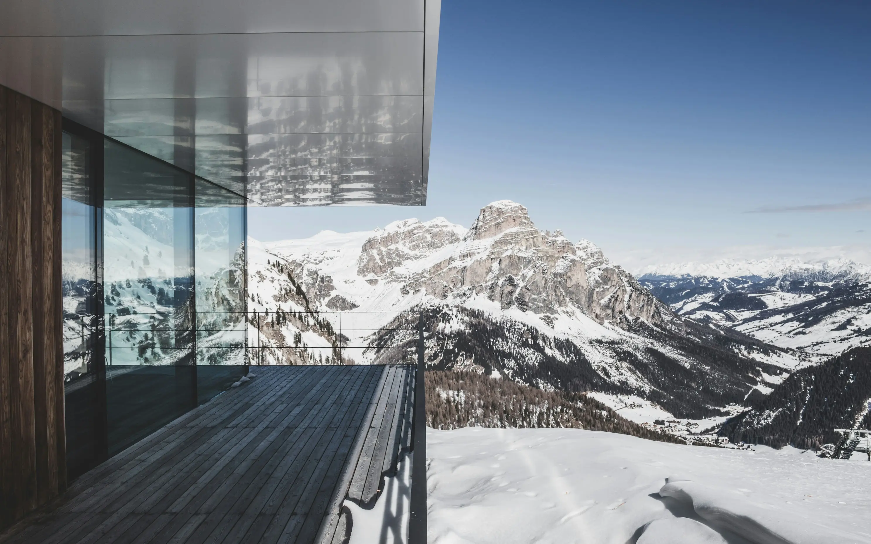 Cette photographie d'architecture capture un moment privilégié sur une terrasse de chalet ou résidence haut de gamme offrant l'une des vues les plus spectaculaires qui soit sur les Alpes italiennes. L'image illustre parfaitement la philosophie architecturale contemporaine de connexion maximale avec le paysage montagnard. Architecture - matériaux et conception: La construction affirme une modernité radicale et sophistiquée : façade entièrement vitrée du sol au plafond avec menuiseries aluminium à profils fins maximisant la transparence, bardage vertical en bois naturel (probablement mélèze ou épicéa) aux teintes grises patinées, terrasse en lames de bois exotique ou composite gris foncé résistant aux conditions climatiques extrêmes, débord de toiture ou casquette en béton brut ou enduit gris clair protégeant la terrasse, garde-corps vitré transparent préservant intégralement la vue panoramique, et réflexions subtiles du paysage dans les vitrages créant des effets de superposition poétiques. La terrasse - espace de vie extérieur: Cet espace extérieur généreux offre une expérience unique : plancher en lames horizontales parfaitement alignées créant des lignes de fuite, orientation plein sud ou sud-ouest capturant lumière et chaleur maximales, protection partielle par le large débord de toiture permettant usage même par temps incertain, garde-corps minimaliste en verre feuilleté sans montants verticaux pour transparence totale, et absence volontaire de mobilier sur cette vue soulignant la pureté architecturale et la prééminence du paysage. Le panorama - les Dolomites: La vue révèle un paysage montagnard parmi les plus spectaculaires d'Europe : massif calcaire caractéristique des Dolomites avec ses tours et falaises verticales emblématiques, sommets enneigés aux formes tourmentées et aux parois stratifiées reconnaissables entre mille, étagement des versants avec forêts de conifères sombres contrastant avec la neige immaculée, vallée habitée visible en contrebas avec hameaux et prairies d'altitude, chaîne de montagnes lointaines s'estompant à l'horizon créant une profondeur infinie, et ciel bleu azur intense typique des belles journées d'hiver en altitude. Lumière et conditions: L'éclairage hivernal sublime l'ensemble : soleil d'hiver bas créant des ombres portées graphiques sur le sol de la terrasse, lumière rasante révélant chaque détail des reliefs rocheux et des formations neigeuses, ciel parfaitement dégagé promettant une journée exceptionnelle, atmosphère cristalline de haute montagne offrant une visibilité exceptionnelle, et reflets du paysage enneigé dans les vitrages amplifiant la sensation d'immersion totale.