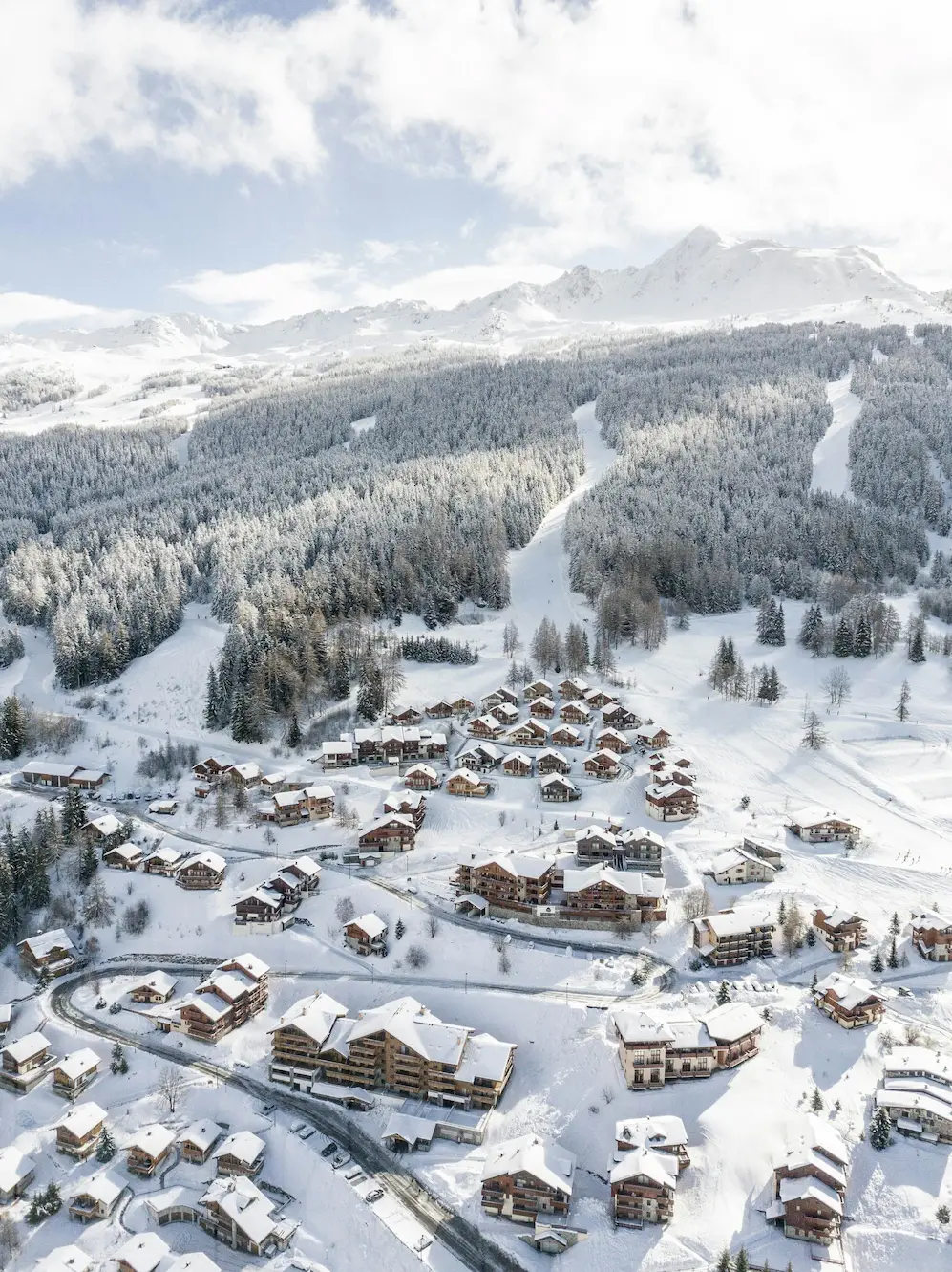 Cette photographie aérienne capturée par drone offre une perspective exceptionnelle sur un village de montagne authentique dans son environnement hivernal immaculé. La vue plongeante révèle l'harmonie parfaite entre habitat traditionnel et nature alpine préservée. Le hameau - organisation et architecture : Le village traditionnel s'organise de manière organique sur un replat naturel : une cinquantaine de chalets et résidences aux toitures généreusement chargées de neige fraîche, architecture alpine authentique avec bardages bois sombre et volumes traditionnels caractéristiques, implantation respectant la topographie naturelle avec constructions suivant les courbes de niveau, voirie enneigée serpentant entre les bâtiments et structurant l'espace villageois, quelques parkings et espaces de stationnement visibles en périphérie, et organisation concentrique autour d'un espace central ou d'une place. On distingue différentes typologies architecturales : chalets individuels traditionnels aux proportions familiales et toitures à forte pente, petits collectifs de standing de 2 à 3 niveaux aux lignes contemporaines mais respectueuses des codes alpins, quelques mazots ou granges anciennes préservées témoignant de l'histoire agricole, et constructions plus récentes parfaitement intégrées au tissu existant. Environnement forestier immédiat : Le village est enchâssé dans un cadre naturel exceptionnel : forêts denses de sapins et d'épicéas enneigés couvrant les versants environnants, arbres givrés créant un camaïeu de blancs immaculés et de verts profonds, clairières et prairies d'altitude ensevelies sous l'épaisse couche neigeuse, quelques arbres isolés (mélèzes dénudés, bouleaux) ponctuant les espaces ouverts, et pistes de ski ou chemins forestiers serpentant entre les arbres en arrière-plan gauche. Domaine skiable et arrière-plan montagneux : Un amphithéâtre de sommets spectaculaires ferme l'horizon : massif enneigé aux formes imposantes culminant probablement au-dessus de 2800-3000 mètres, versants alternant zones rocheuses abruptes et pentes enneigées skiables, pistes damées visibles serpentant dans la forêt et s'ouvrant sur les combes d'altitude, quelques installations de remontées mécaniques discrètement intégrées, et ciel partiellement nuageux créant des jeux de lumière dramatiques sur les sommets. Conditions et atmosphère : L'image capture un moment privilégié : enneigement optimal témoignant de récentes chutes de neige abondantes, lumière hivernale douce et homogène typique d'une matinée ou après-midi d'hiver, absence quasi totale de traces dans la neige suggérant un lendemain de chute ou un moment de calme, atmosphère de quiétude et de sérénité absolue, et sentiment d'isolement préservé au cœur d'un environnement naturel exceptionnel. Cette vue identique à une précédente confirme le caractère exceptionnel du site.