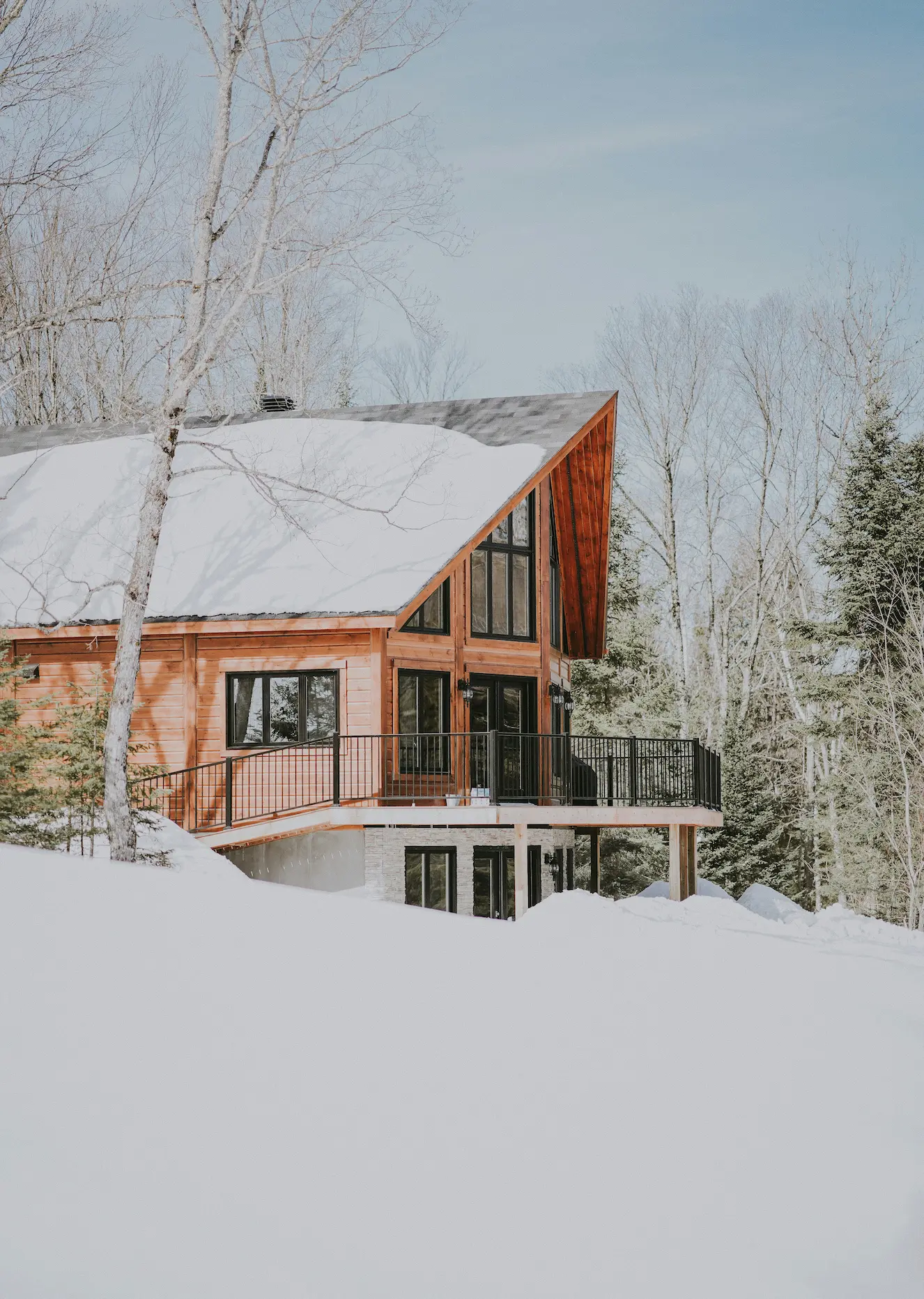 Voici une description de cette image : Description courte : Chalet contemporain en bois implanté dans une forêt enneigée, avec architecture moderne à pignon vitré, large terrasse sur pilotis et toiture recouverte de neige épaisse. Description détaillée : Cette photographie hivernale présente un chalet individuel d'architecture contemporaine parfaitement intégré dans un environnement forestier de montagne. L'image capture l'harmonie entre construction moderne et nature enneigée dans une atmosphère paisible et lumineuse. Architecture : Le bâtiment affirme une identité alpine contemporaine avec des volumes géométriques francs. La façade principale présente un imposant pignon vitré sur deux niveaux inondant l'intérieur de lumière naturelle, encadré de menuiseries bois sombre créant un rythme vertical élégant. Les façades latérales sont entièrement habillées de bardage bois horizontal dans des tons miel chaleureux qui contrasteront magnifiquement avec le blanc de la neige. La toiture à double pente largement débordante est couverte d'une épaisse couche de neige fraîche, témoignant des conditions hivernales rigoureuses que le bâtiment supporte sans difficulté. Espaces extérieurs : Une généreuse terrasse sur pilotis en bois clair s'étend sur toute la largeur de la façade, créant un espace extérieur suspendu au-dessus de la pente naturelle. Le garde-corps métallique à barreaux verticaux noirs apporte une touche contemporaine tout en assurant sécurité et transparence. Cette terrasse accessible depuis le niveau de vie principal permet de profiter de l'extérieur même en plein hiver. Le soubassement en béton gris visible sous la terrasse ancre solidement la construction dans le terrain en pente. Environnement : Le chalet est immergé dans une forêt mixte enneigée composée de bouleaux aux troncs blancs caractéristiques (dénudés en hiver) et de conifères persistants givrés. L'épaisse couche de neige immaculée au sol crée un tapis blanc uniforme soulignant la pureté de l'environnement. L'arrière-plan légèrement brumeux suggère la profondeur de la forêt et renforce l'atmosphère feutrée typique des journées d'hiver en montagne. Le ciel bleu pâle et la lumière douce naturelle créent une ambiance sereine. Atmosphère : L'ensemble dégage une sensation de quiétude, d'intimité et de connexion avec la nature, tout en affirmant un confort moderne. Le chalet apparaît comme un refuge chaleureux au cœur de l'hiver, parfaitement équipé pour profiter des joies de la saison froide.