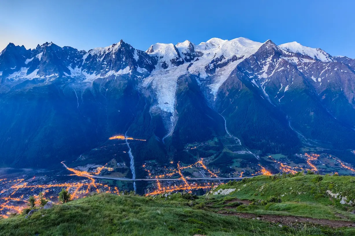 Cette photographie panoramique exceptionnelle capture l'instant magique de la tombée de la nuit dans une vallée alpine majeure, probablement la vallée de Chamonix face au massif du Mont-Blanc. L'image révèle la cohabitation harmonieuse entre présence humaine et nature grandiose. Au premier plan : Des alpages verdoyants typiques de la moyenne montagne, avec herbes grasses et quelques arbustes isolés. Un sentier de randonnée serpente sur la pente, témoin de la fréquentation estivale des lieux. La végétation luxuriante contraste avec la minéralité des sommets. Au second plan : La vallée s'illumine progressivement à l'heure bleue, révélant un réseau urbain étendu structuré par l'éclairage public orange caractéristique. Les routes principales dessinent des lignes lumineuses rectilignes traversant la vallée, tandis que les hameaux et villages créent des constellations de points orangés dispersés sur les replats et les coteaux. Cette urbanisation linéaire typique des vallées alpines souligne la géographie contrainte du développement humain en montagne. Au troisième plan : Les versants boisés plongent dans l'ombre crépusculaire, révélant les reliefs escarpés et les couloirs d'avalanche qui strient les pentes abruptes. L'étagement de la végétation (forêts de conifères, puis zone de combat, puis pelouses alpines) est subtilement visible malgré la pénombre. À l'horizon : Le massif du Mont-Blanc déploie toute sa majesté sous les dernières lueurs du jour. Les sommets enneigés et les glaciers suspendus captent encore la lumière, créant un dégradé subtil du blanc éclatant au bleu profond. Les arêtes acérées, les calottes glaciaires et les faces verticales se détachent sur le ciel bleu intense du crépuscule alpin. La ligne de crête dentelée révèle l'altitude exceptionnelle de ce massif mythique culminant à plus de 4800 mètres. L'ensemble crée une composition saisissante entre l'intimité de la vie humaine illuminée dans la vallée et la puissance intemporelle des géants de glace.