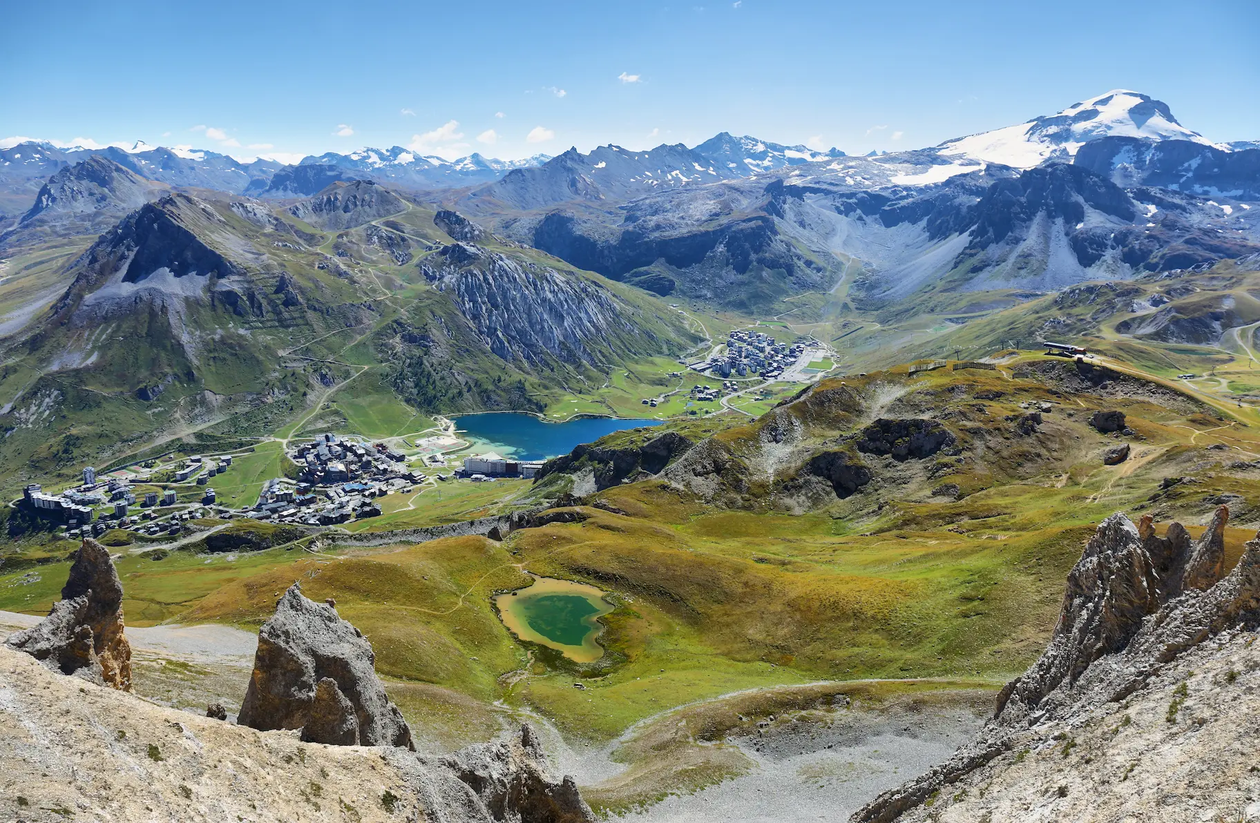 Cette photographie aérienne spectaculaire capture l'essence d'une station de sports d'hiver de haute altitude durant la saison estivale. Prise depuis un sommet environnant, elle offre une perspective unique sur l'organisation urbaine et l'environnement naturel exceptionnel. Au premier plan : Des formations rocheuses ériques et minérales caractéristiques de la haute montagne, avec affleurements de schistes gris et éboulis. Un petit lac d'altitude aux eaux turquoise émeraude témoigne de la richesse hydrologique du site. Au second plan : La station se déploie en deux secteurs distincts reliés par la voirie. À gauche, un premier hameau de constructions alpines compactes ; au centre-gauche, un lac de barrage ou naturel aux eaux d'un bleu profond intense bordé de prairies d'altitude ; et légèrement en arrière, un second secteur d'habitations plus densément construit. L'architecture respecte les codes alpins avec toitures pentues et volumes maîtrisés s'intégrant dans la topographie. Au troisième plan : Les versants montagneux révèlent une mosaïque paysagère typique : prairies d'altitude aux teintes vert tendre ponctuées de zones d'éboulis gris, crêtes rocheuses acérées, couloirs d'avalanche dénudés, et sentiers de randonnée serpentant sur les pentes. La limite entre étage alpin herbeux et étage subalpin rocheux est clairement visible. À l'horizon : Un amphithéâtre majestueux de sommets enneigés éternels se déployant à perte de vue, avec glaciers suspendus, calottes sommitales et arêtes déchiquetées. Le ciel bleu azur parsemé de quelques nuages cotonneux sublime la pureté de l'atmosphère d'altitude. L'ensemble illustre parfaitement l'équilibre entre développement touristique maîtrisé et préservation d'un environnement alpin d'exception.