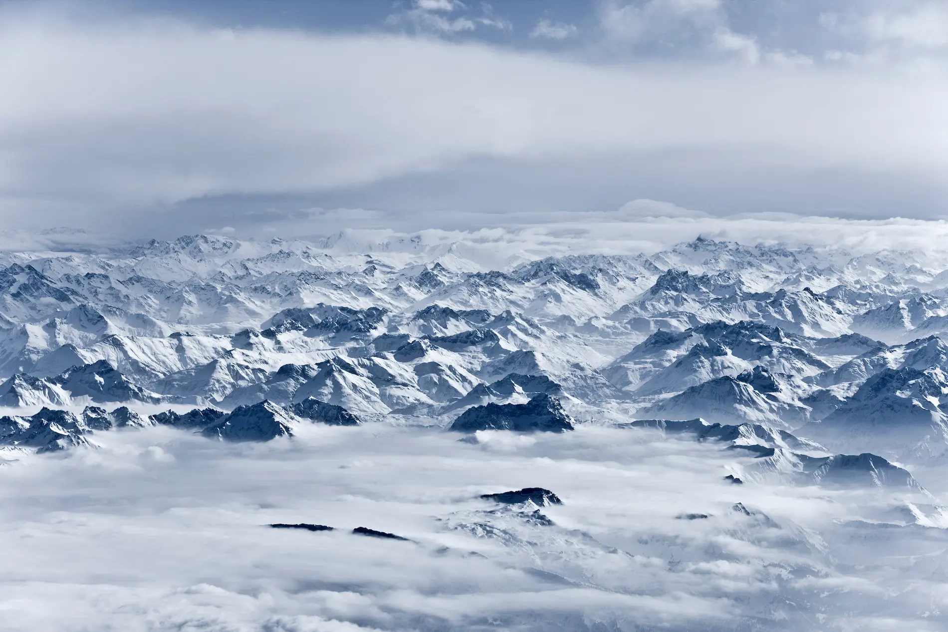Cette photographie aérienne ou prise depuis un sommet d'altitude capture l'essence même de la haute montagne alpine dans toute sa splendeur hivernale. L'image révèle un paysage grandiose et infini de massifs montagneux enneigés s'étageant à perte de vue. Le premier plan montre quelques sommets rocheux sombres émergeant d'une épaisse couche de nuages bas, créant un effet de mer de brume ouateuse qui enveloppe les vallées invisibles en contrebas. Cette inversion thermique, phénomène typique de la montagne hivernale, isole visuellement les sommets et accentue leur caractère inaccessible et majestueux. Le second plan dévoile une succession de chaînes montagneuses parallèles créant une profondeur spectaculaire : crêtes acérées, versants raides striés par les couloirs d'avalanche, faces nord ombragées contrastant avec les versants sud plus lumineux, glaciers suspendus et névés persistants. L'étagement des plans crée un dégradé subtil de tons bleutés caractéristique de la perspective atmosphérique en haute altitude. L'arrière-plan se perd dans un horizon brumeux où les sommets les plus lointains se fondent dans le ciel nuageux gris-bleu, suggérant l'immensité du massif alpin. La lumière diffuse et les nuages menaçants en altitude renforcent l'atmosphère dramatique et la puissance des éléments naturels. Cette vue incarne parfaitement l'environnement exceptionnel des stations de sports d'hiver de haute altitude et la majesté des Alpes françaises.